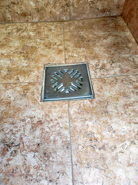 Square metal floor drain set into textured stone tile bathroom floor. Plumbing fixture for shower drainage and water management. Home maintenance and interior construction detail