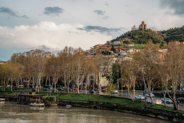 Kura River embankment with plane trees and hillside houses beneath a hilltop church in Tbilisi, Georgia © Puripat