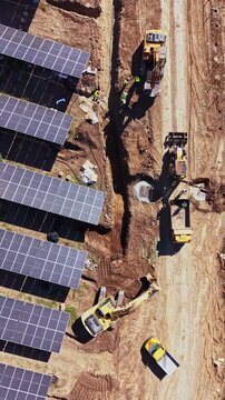 An aerial view shows excavators and trucks working beside a trench at a solar farm construction site, with rows of panels under the sun.