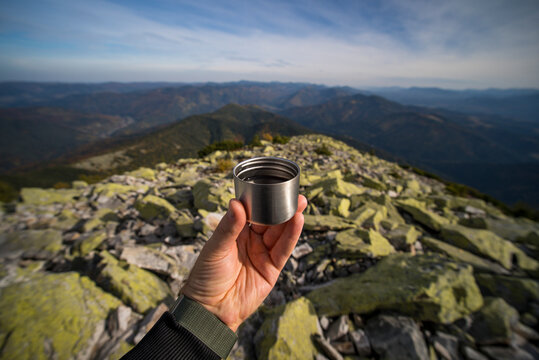 Male hand holding a steel thermos cup on a mountain peak against a panoramic view of mountain ranges and blue sky. Hiking, outdoor adventure, travel, hot drink on top, freedom lifestyle concept.