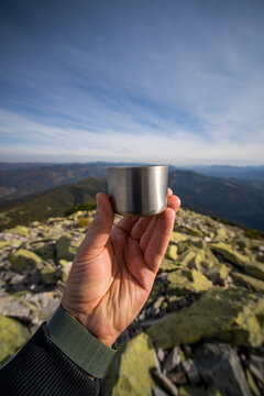 Male hand holding a steel thermos cup on a mountain peak against a panoramic view of mountain ranges and blue sky. Hiking, outdoor adventure, travel, hot drink on top, freedom lifestyle concept.