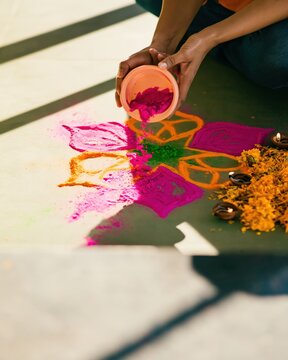 Close-up of hands pouring vivid magenta powder into a colorful rangoli design with marigold petals and clay oil lamps
