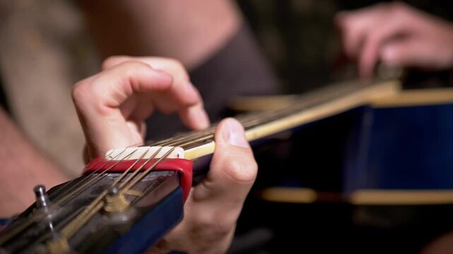 Hand frets acoustic guitar strings against a wooden neck fitted with a red capo. The detailed macro perspective serves tutorials, songwriting sequences, and musical scoring.