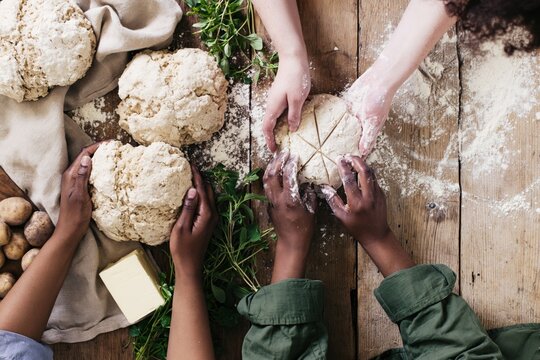 Overhead view of diverse hands shaping rustic scored bread loaves on a flour dusted wooden table with potatoes, butter and herbs