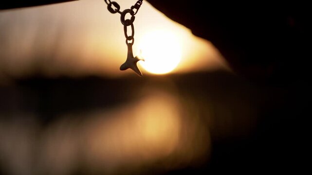 A small bird pendant dangles from a delicate chain bracelet during sunset. The metallic silhouette shines in soft counterlight with blurred water bokeh and static macro framing.