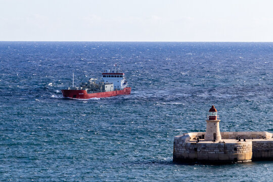 Valletta, Malta - October 14th, 2026 Red Cargo Ship Passing A Stone Lighthouse At Sea Near A Harbor Breakwater On A Windy Day