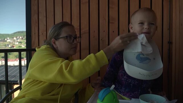 A mom in a yellow hoodie wipes her child's mouth and hands with a napkin after breakfast. the child, wearing a bib, stands up and reaches toward camera, calm family morning terrace country house.