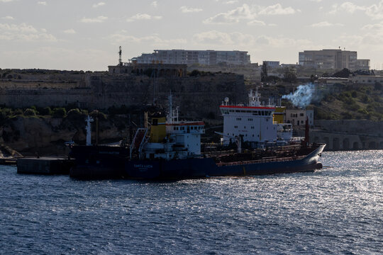 Valletta, Malta - October 14th, 2026 Cargo Ships Docked at Industrial Port Beside Fortified Coastline With Sunlit Sea