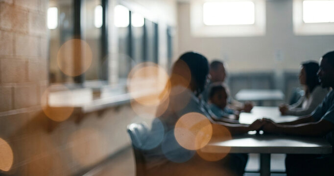A group of individuals sits around a table in a subdued environment, holding hands and engaging in a moment of connection. The soft focus creates a contemplative mood.