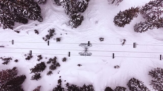 Chairlift cables over snow covered forest and ski track. Cableway chairs, steel support tower, snowbound pine tree tops and ski tracks in snow.
