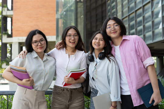 Asian university students stand together outdoors with books, headphones, and a laptop, showing friendship, student lifestyle, education, campus community, and relaxed university life.