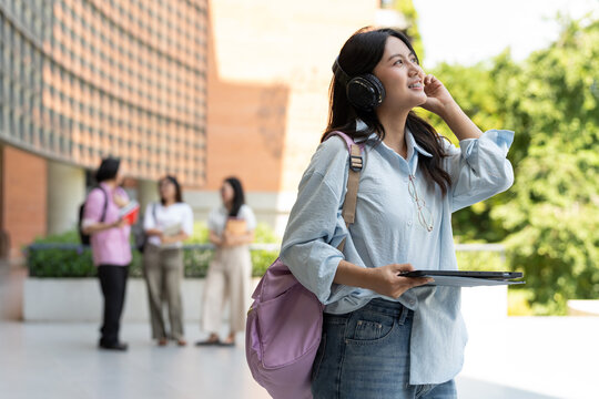Asian university student wearing headphones and carrying a tablet walks outdoors on campus, combining digital learning, student lifestyle, technology, and everyday education in a modern setting.