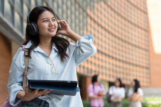 Asian university student listens through headphones while holding a tablet outdoors, with classmates behind her, creating a modern student lifestyle scene for education, technology, and youth.