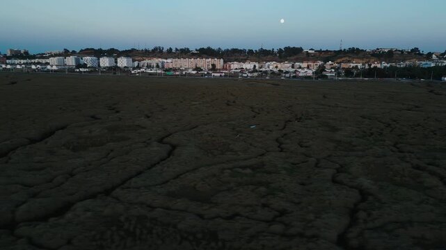 Aerial drone shot gliding forward over a vast cracked mudflat toward the distant illuminated Huelva skyline at twilight with the moon rising in an urban expansion and environmental change concept.