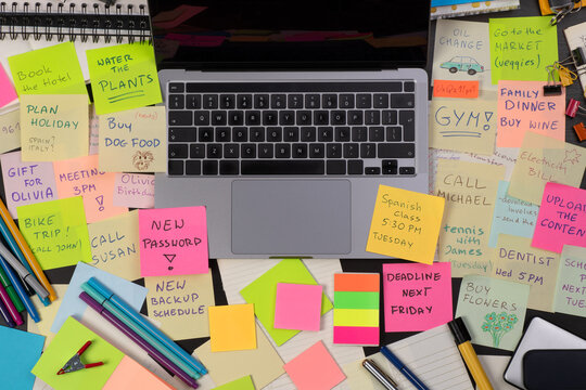 Messy office desk of a very busy person with an open laptop, covered in colorful sticky notes, top view
