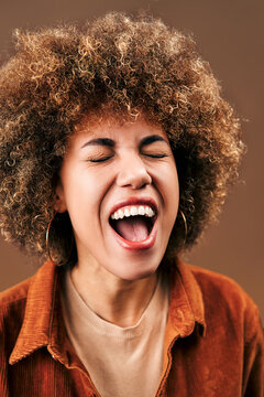 Portrait of a joyful woman with voluminous afro hair, large hoop earrings screaming with her eyes closed and mouth wide open.Expression of excitement, euphoria, liberation, and unrestrained emotion.