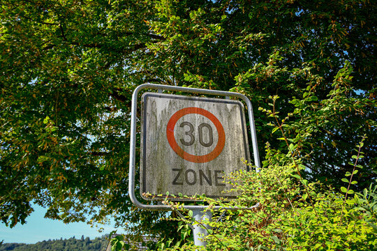 A weathered mossy German thirty-kilometre zone traffic sign stands among lush green summer tree foliage