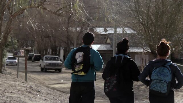 Three High School Students Standing with Backpacks Looking at Dirt Street in Chos Malal  