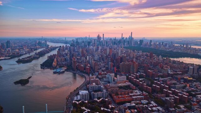 Aerial view of Manhattan buildings and East River during sunset. Dense rows of urban structures extend toward a wide river under a colorful sky.