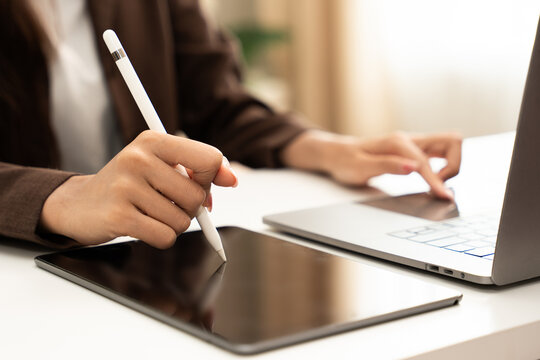 Digital workspace concept. Close-up of a woman using a stylus on a tablet while operating a laptop at a desk indoors in natural light. Tablet, stylus, laptop, technology, multitasking.