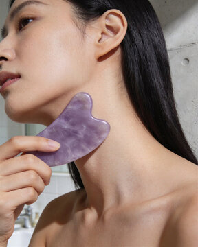 Young Asian woman demonstrating gua sha scraping neck technique with purple amethyst crystal tool in bathroom for skin firming.