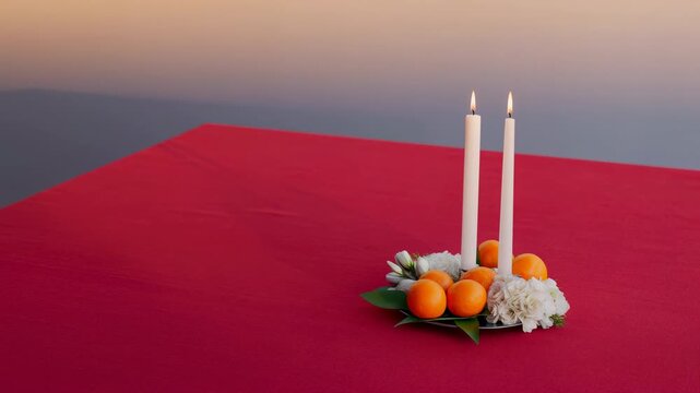 Two white taper candles burn among oranges and white flowers on red tablecloth as warm directional light fades and long diagonal shadows soften against plain beige wall