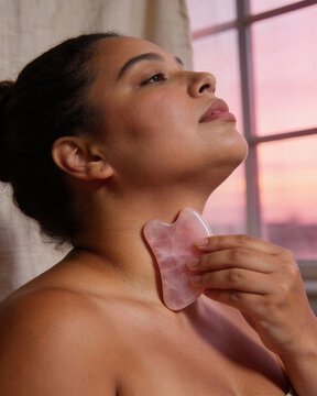 Woman practicing gua sha scraping neck technique with rose quartz crystal tool near window at sunset for lymphatic drainage