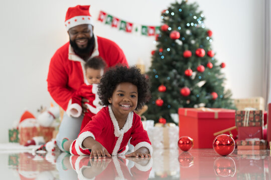 African American family, father, little son and baby boy in red Santa Claus costume spending time together and celebrates Christmas in festive living room near decorated tree and festive decorations