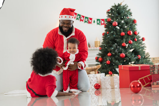 African American family, father, little son and baby boy in red Santa Claus costume spending time together and celebrates Christmas in festive living room near decorated tree and festive decorations