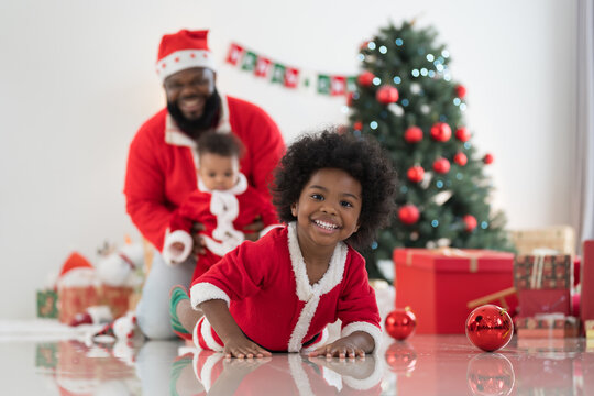 African American family, father, little son and baby boy in red Santa Claus costume spending time together and celebrates Christmas in festive living room near decorated tree and festive decorations