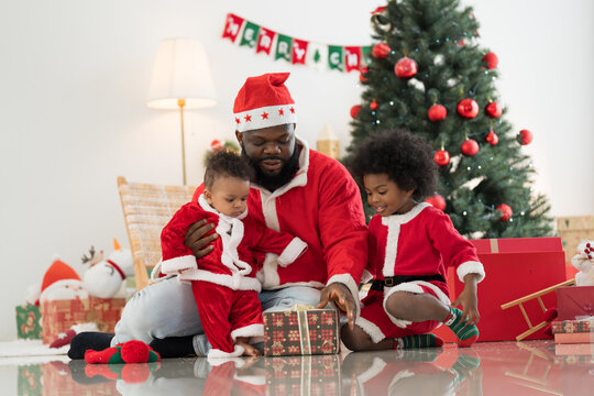 African American family, father, little son and baby boy in red Santa Claus costume spending time together and celebrates Christmas in festive living room near decorated tree and festive decorations