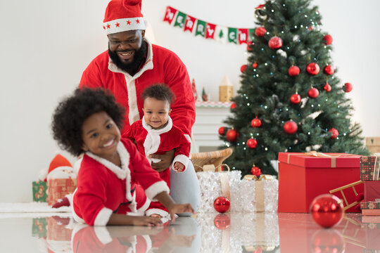 African American family, father, little son and baby boy in red Santa Claus costume spending time together and celebrates Christmas in festive living room near decorated tree and festive decorations