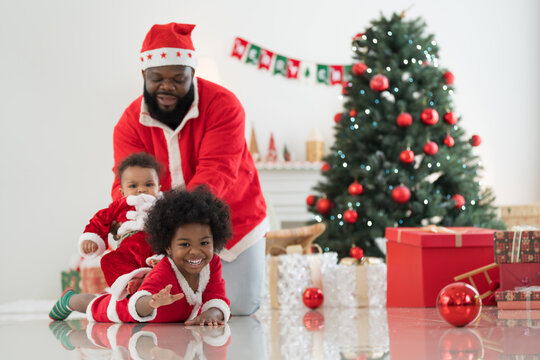 African American family, father, little son and baby boy in red Santa Claus costume spending time together and celebrates Christmas in festive living room near decorated tree and festive decorations