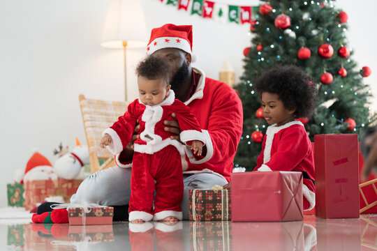 African American family, father, little son and baby boy in red Santa Claus costume spending time together and celebrates Christmas in festive living room near decorated tree and festive decorations