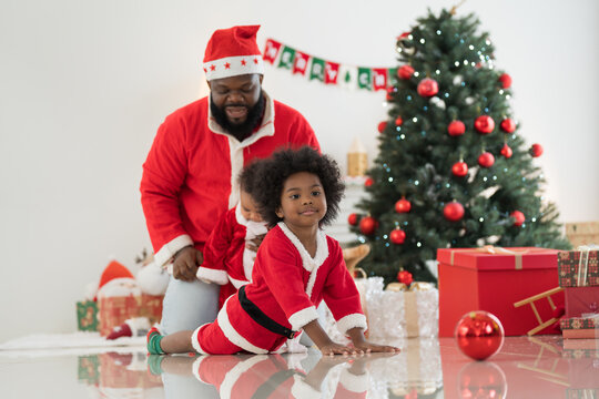 African American family, father, little son and baby boy in red Santa Claus costume spending time together and celebrates Christmas in festive living room near decorated tree and festive decorations