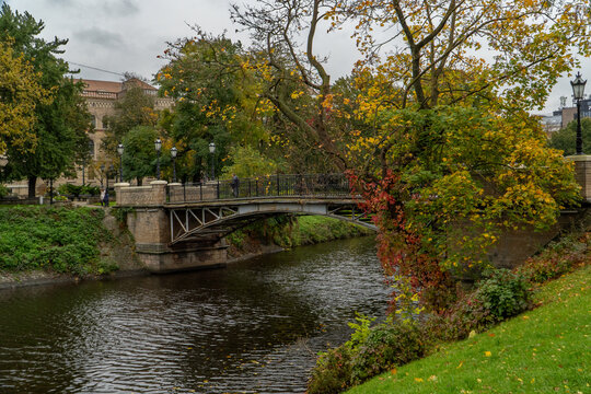 Autumn Park Bridge Over River With Trees And Historic City Buildings On A Cloudy Day