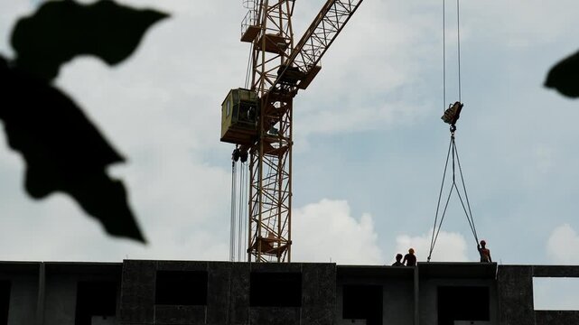 Tower crane operates above silhouette construction workers standing atop unfinished concrete building under cloudy sky, as framing leaves create natural foreground border during daytime work.