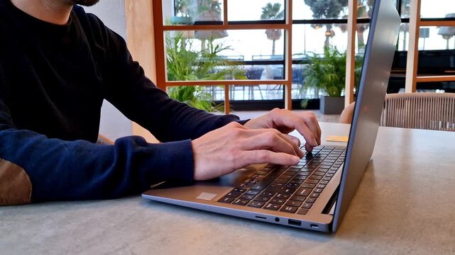 Person working on a laptop at a table in a modern cafe or workspace with large windows and a bright interior