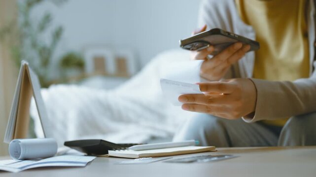 Expense tracking with a smartphone and receipt, hands comparing the receipt beside a notebook on a home table, keeping the smartphone central as receipt review accompanies a mobile budgeting routine.