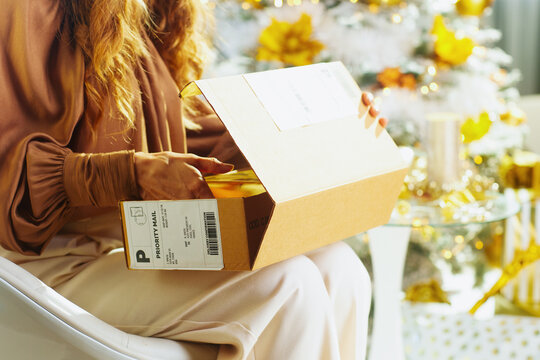 A woman with wavy brown hair sits by a festive Christmas tree, eagerly opening a cardboard box with a shipping label. The scene captures the anticipation of receiving a holiday gift.