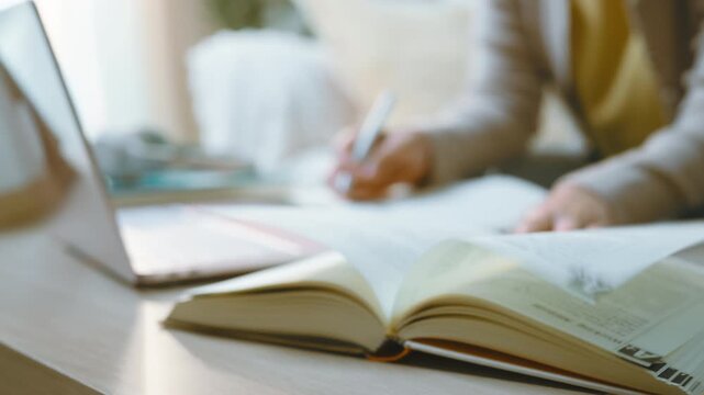 Home study centers on a book in the foreground as a person continues writing notes beside an open laptop, with the book anchoring a quiet tabletop view of home study during revision.