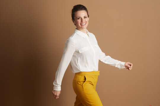 A mid-adult businesswoman, dressed in a crisp white shirt and yellow pants, walks with a confident smile, looking at the camera, symbolizing career progression and success.