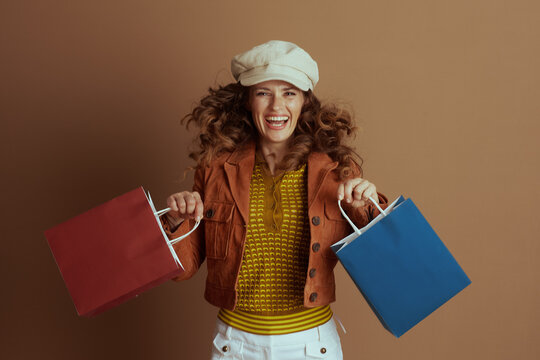 A joyful woman with long curly hair, dressed in a rust-colored jacket and cap, is captured mid-jump, enthusiastically holding two shopping bags against a plain brown background.