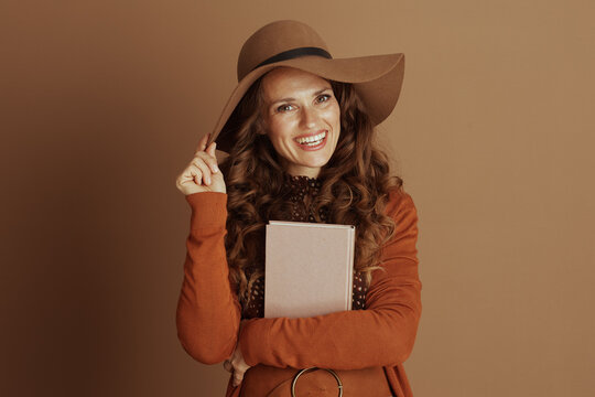 A cheerful woman with curly brown hair, dressed in autumnal fashion with a wide-brimmed hat and orange cardigan, confidently holds a pink book against her chest on a warm brown background.