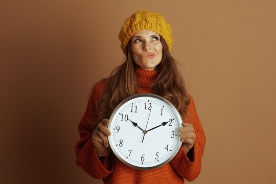 A woman in yellow beanie and orange turtleneck sweater holds large wall clock, looking upwards with thoughtful expression. The scene conveys contemplation about time, scheduling, or seasonal changes