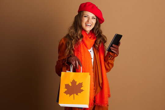 A smiling woman in red beret and orange scarf holds orange shopping bag and smartphone, against plain brown background. Her orange jacket and brown gloves convey cheerful, stylish autumnal mood