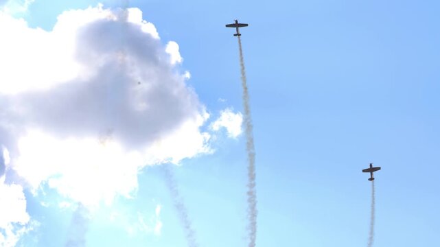 Aerobatic propeller planes perform loops and vertical maneuvers leaving white smoke trails against blue sky and clouds, sun rays, low angle view