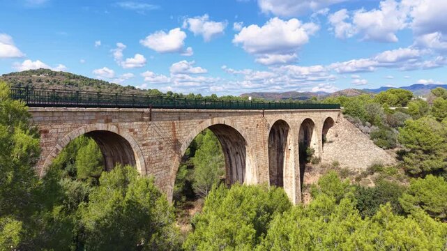 Old stone arched viaduct spanning a wooded ravine with green metal railing, surrounded by Mediterranean pine trees and rolling hills under cumulus clouds