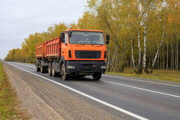 Orange dump truck with a trailer on a forest road. Construction machinery moving along a route through an autumn forest. Heavy-duty vehicle with a dump body, designed for transporting loose materials © yaviki
