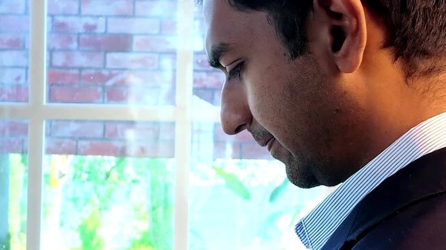 Young Indian businessman in a formal suit attending a video conference on his laptop while standing near a window in a modern office, representing corporate communication and remote business meetings.